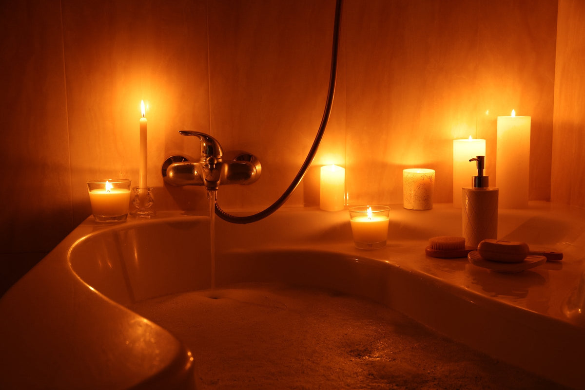 A bath filling with water in a bathroom lit by burning candles, with soap and a brush at the edge