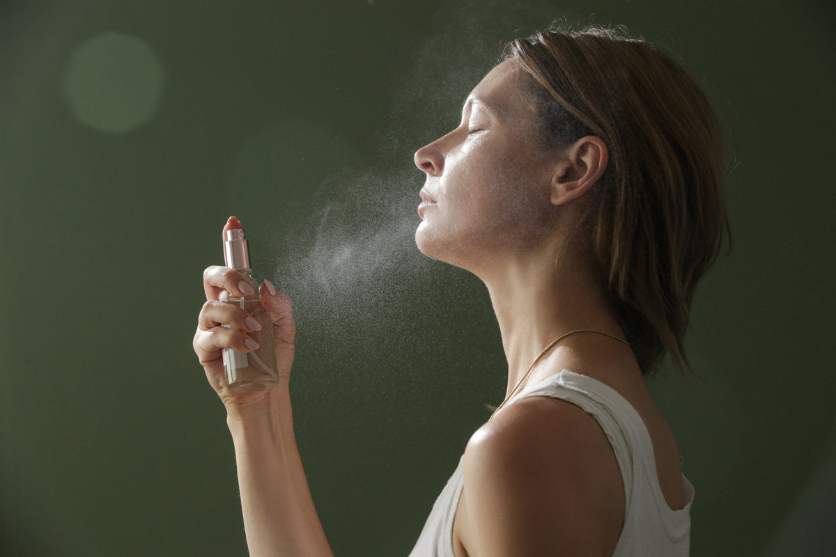 A woman with her eyes closed spraying a fine facial mist onto her face, against a dark green background