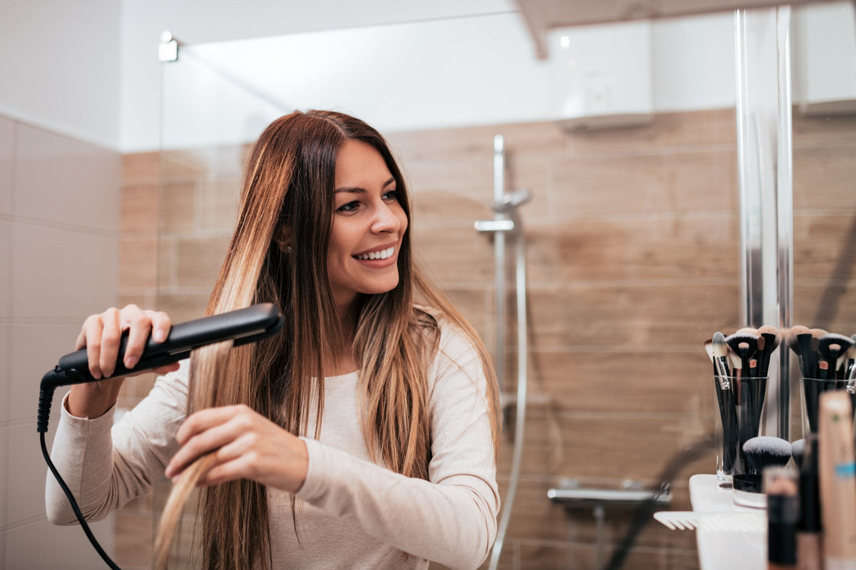 A smiling woman straightening her long hair with a flat iron at a bathroom mirror