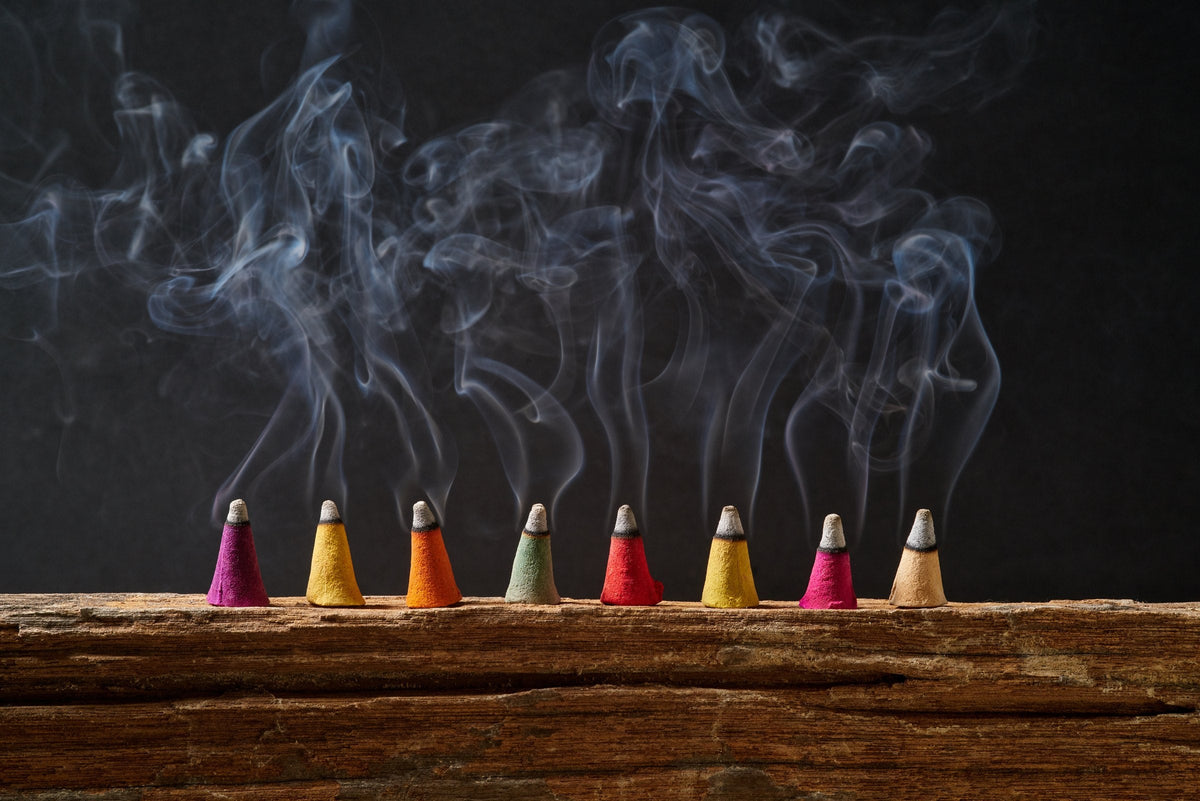 A row of coloured incense cones on old wood, with smoke curling up against a black background