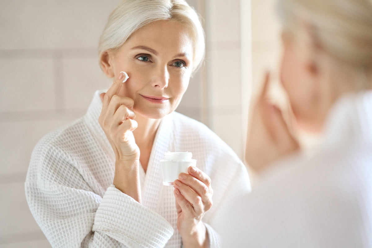 A woman around fifty in a white robe applying face cream
